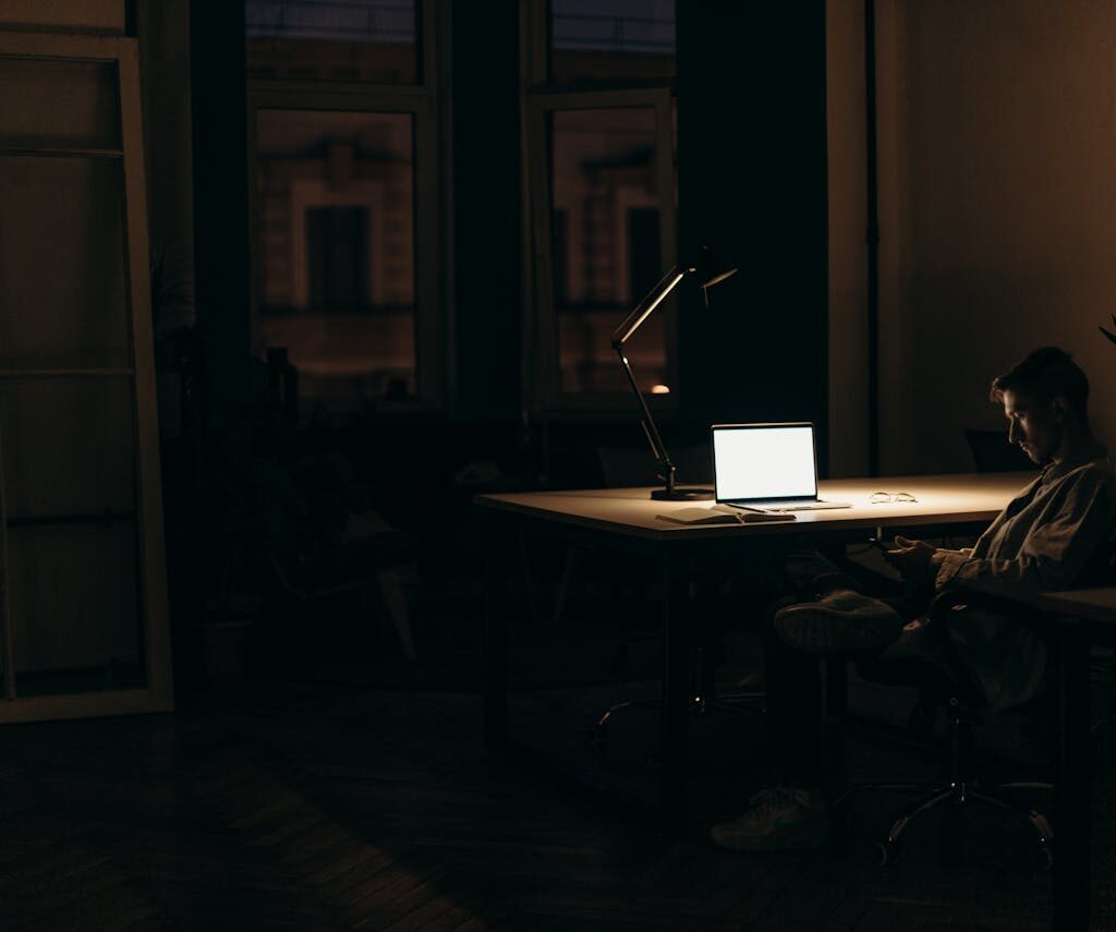 A man working late at night in a dimly lit office, with a bright laptop screen.